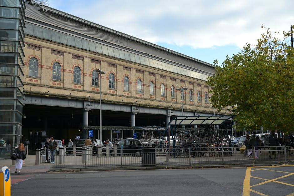 The image shows the exterior of Kilburn Park Station building, featuring an old brick facade with arched windows and decorative red brick accents, situated above an open parking area. In the foregroun